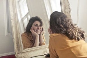 stock image of young woman looking at herself in the mirror smiling with both palms on chin wearing a yellow hoodie