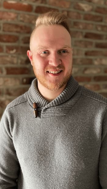 Therapist Tomas Clarke standing indoors by a brick wall wearing a gray sweater